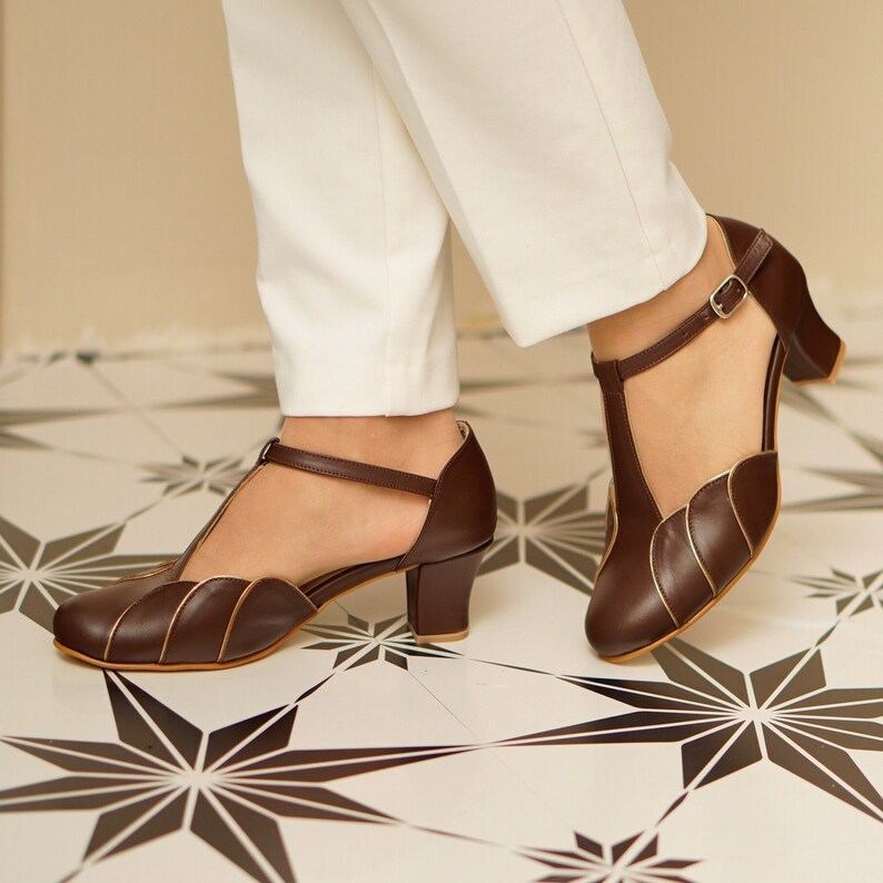 May include: Brown leather heeled shoes with a T-strap and gold trim. The shoes have a scalloped design on the toe and a small heel.