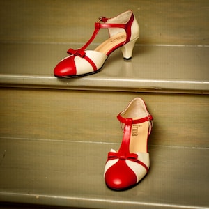 Pu&ograve; includere: Un paio di scarpe con tacco rosso e bianco con un design a T. Le scarpe hanno un dettaglio a fiocco sulla punta e il nome del marchio "Irma" &egrave; stampato sulla soletta.