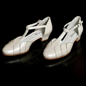 May include: A pair of white leather t-strap shoes with a low heel. The shoes have a decorative design on the toe and are labeled "Irma" on the insole.