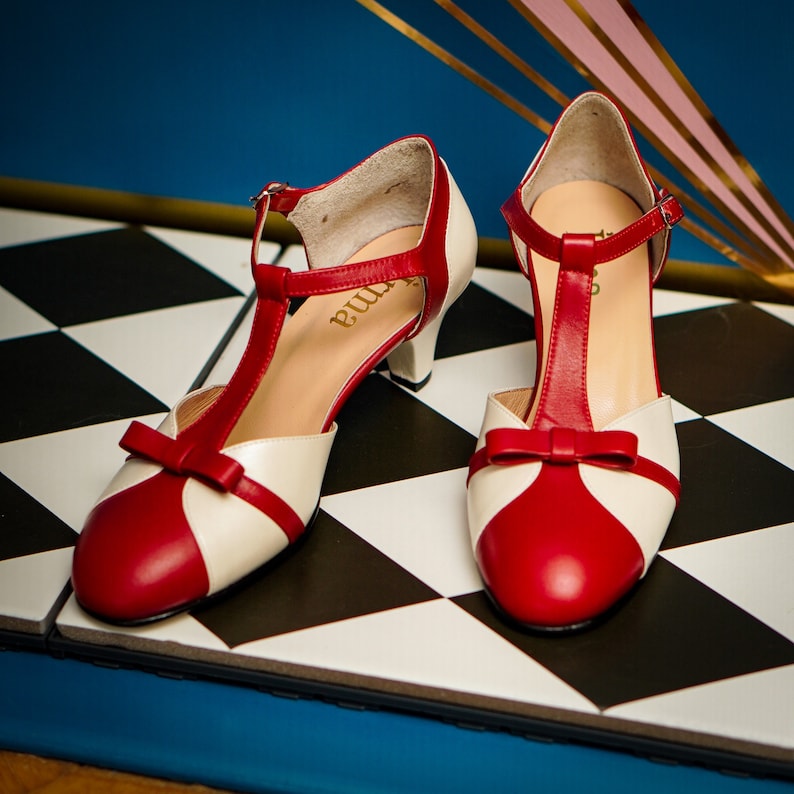 Pu&ograve; includere: Un paio di scarpe con tacco rosso e bianco con cinturino a T e fiocco sulla punta. Le scarpe sono su una superficie a scacchi bianchi e neri.