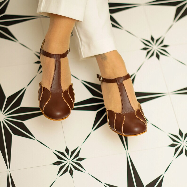 May include: A pair of brown leather heeled shoes with a t-strap design and a scalloped edge. The shoes have a light brown trim and are worn on a white and black tiled floor.