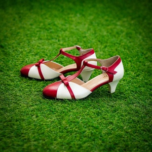 Pu&ograve; includere: Un paio di scarpe con tacco bianco e rosso con cinturino a T e dettaglio a fiocco. Le scarpe sono su una superficie erbosa verde.