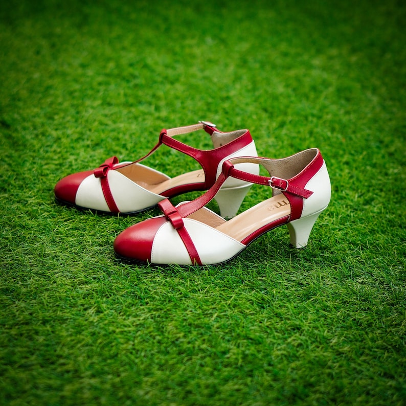 Pu&ograve; includere: Un paio di scarpe con tacco bianco e rosso con cinturino a T e dettaglio a fiocco. Le scarpe sono su una superficie erbosa verde.