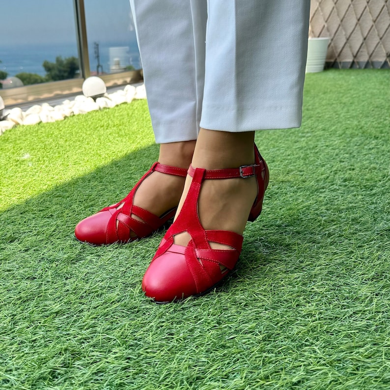 May include: Red leather heeled sandals with a T-strap and cut-out design. The sandals are on a green grassy surface.