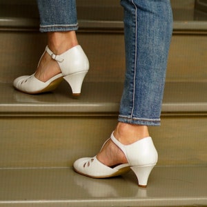 May include: A pair of white heeled shoes with ankle straps. The shoes are worn by a person wearing blue jeans and standing on a set of stairs.