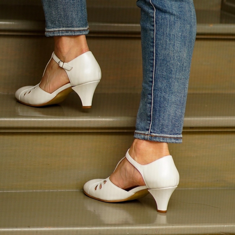 May include: A pair of white heeled shoes with ankle straps. The shoes are worn by a person wearing blue jeans and standing on a set of stairs.