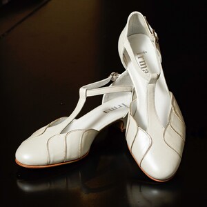 May include: A pair of off-white leather heeled shoes with a T-strap design. The shoes have a scalloped detail on the front and a small heel. The brand name "Irma" is visible on the inside of the shoes.