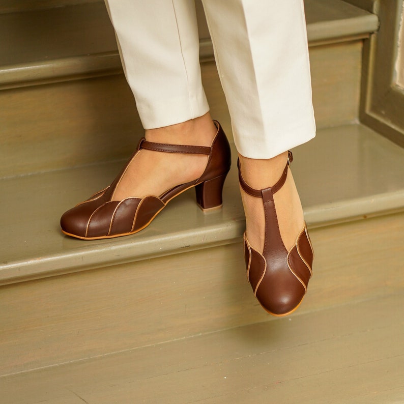 May include: Brown leather heeled shoes with a T-strap and a decorative scalloped design. The shoes have a low block heel.