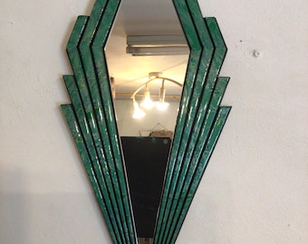 May include: Art Deco style mirror with a geometric design. The mirror has a diamond shape with a green and black frame. The frame features a series of angled lines radiating from the center, creating a fan-like effect. The mirror reflects the surrounding environment.
