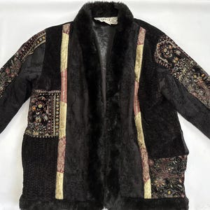 May include: A black jacket featuring a mix of textures and patterns, including solid black faux fur panels, gold-toned fabric strips, and sections with a dark floral paisley print. The garment has a long-sleeve design with a plush faux fur collar.