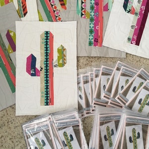 May include: A collection of fabric squares with a cactus design in various colors and patterns. The squares are arranged in a grid pattern on a white surface. There are also several packages of fabric squares with a cactus design in the same colors and patterns. The packages are labeled "Sonoran Saguaro".