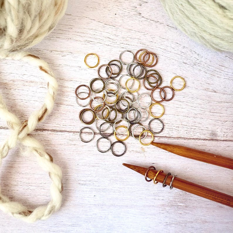 May include: A collection of assorted metal stitch markers in gold, silver, and bronze tones, scattered on a light wood surface. Two wooden knitting needles are also present, with stitch markers attached. Yarn balls are visible in the background, suggesting a knitting or crochet theme.
