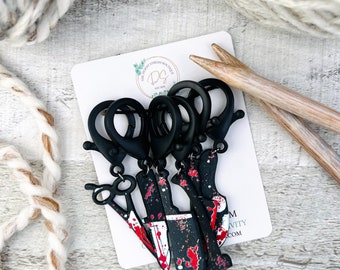 May include: Five black metal stitch markers with red and black patterned charms shaped like knives and scissors. The charms are attached to the stitch markers with silver rings. The stitch markers are on a white card with the text "The Craft Stream Boutique" and a logo.