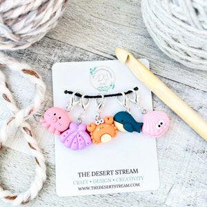 May include: Set of five colorful sea creature stitch markers. The markers are made of polymer clay and feature an octopus, a seashell, a crab, a whale, and a clam. The markers are attached to silver-toned clips. The image also includes a wooden crochet hook and a ball of yarn.