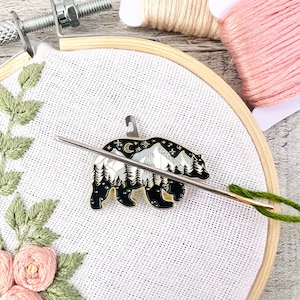 May include: A black bear enamel pin with a white mountain scene and gold accents. The pin is on a white fabric background with a needle and thread.