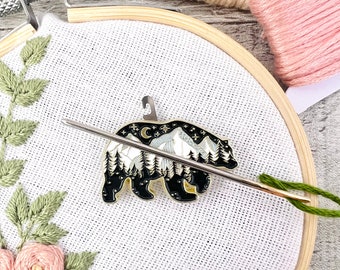 May include: A black bear enamel pin featuring a white mountain scene and gold accents. The pin is displayed on a white fabric background with a needle and thread.