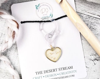May include: A clear plastic stitch marker with a gold heart charm containing white glitter. The stitch marker is attached to a black cord and a clear plastic clip. The stitch marker is on a white card with the text "THE DESERT STREAM" and "CRAFT • DESIGN • CREATIVITY" and "WWW.THEDESERTSTREAM.COM".