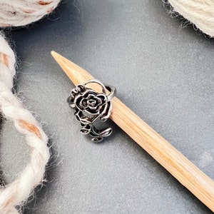 May include: Silver rose stitch marker with a detailed design. The marker is on a wooden knitting needle.