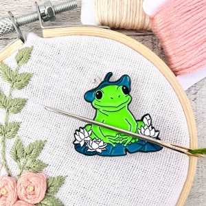 May include: Green frog wearing a blue hat and sitting on a lily pad with white flowers. The frog is a metal pin.