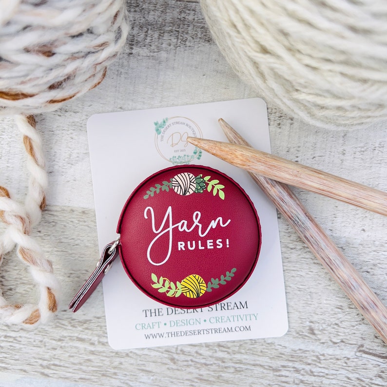 Pu&ograve; includere: Un metro a nastro rotondo bordeaux con la scritta "Yarn Rules!" e decorazioni di filato e foglie. L'immagine include gomitoli di lana, ferri da maglia e una scheda con il logo e il sito web del negozio.