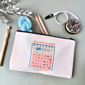 May include: A white canvas pouch with a black zipper, featuring a colorful crochet schedule design. Inside are knitting needles, a ball of blue yarn, and a measuring tape. A wooden crochet hook is also visible.