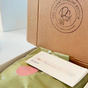 May include: A brown cardboard box with a circular logo that reads "The Desert Stream Stitch." Inside, a package wrapped in green tissue paper and a white card with text that says "Thank you for your order!"