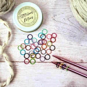 Peut inclure: Des marqueurs de mailles colorés et des aiguilles à tricoter sont disposés sur une surface en bois. Une petite boîte avec le texte "The Desert Stream Stitch" est également présente. Les marqueurs de mailles sont de différentes couleurs, dont le rose, le bleu et le vert.
