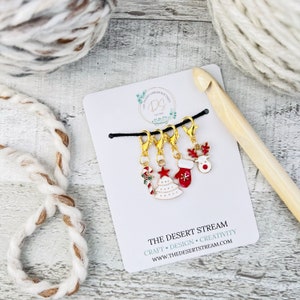 May include: A set of gold-colored Christmas-themed stitch markers with red, white, and green enamel details. The markers include a candy cane, Christmas tree, mitten, and reindeer charm. The card reads "The Desert Stream Boutique".