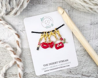 May include: Set of five gold stitch markers with red and white enamel charms. The charms include a lipstick, a heart with the word "love", a purse, and two lips. The stitch markers are on a white card with the text "The Desert Stream" and a website address.
