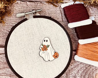 May include: A white ghost holding a bouquet of orange flowers and a small orange pumpkin with a jack-o'-lantern face. The ghost is pinned to a white fabric stretched over a black embroidery hoop.