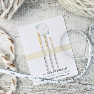 May include: A white card with the text "The Desert Stream Boutique" and "The Desert Stream" with the words "Craft, Design, Creativity" and "thedesertstream.com". Three needles are displayed on the card, two silver and one gold.