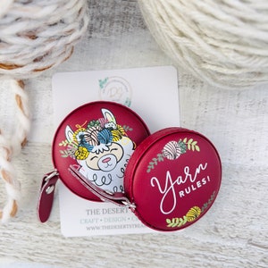Pu&ograve; includere: Due metri a nastro rotondi bordeaux. Uno presenta un lama con filato e fiori, l'altro dice "Yarn Rules!" in corsivo bianco. Una carta bianca con testo &egrave; dietro i metri a nastro. Matasse di filato sullo sfondo.