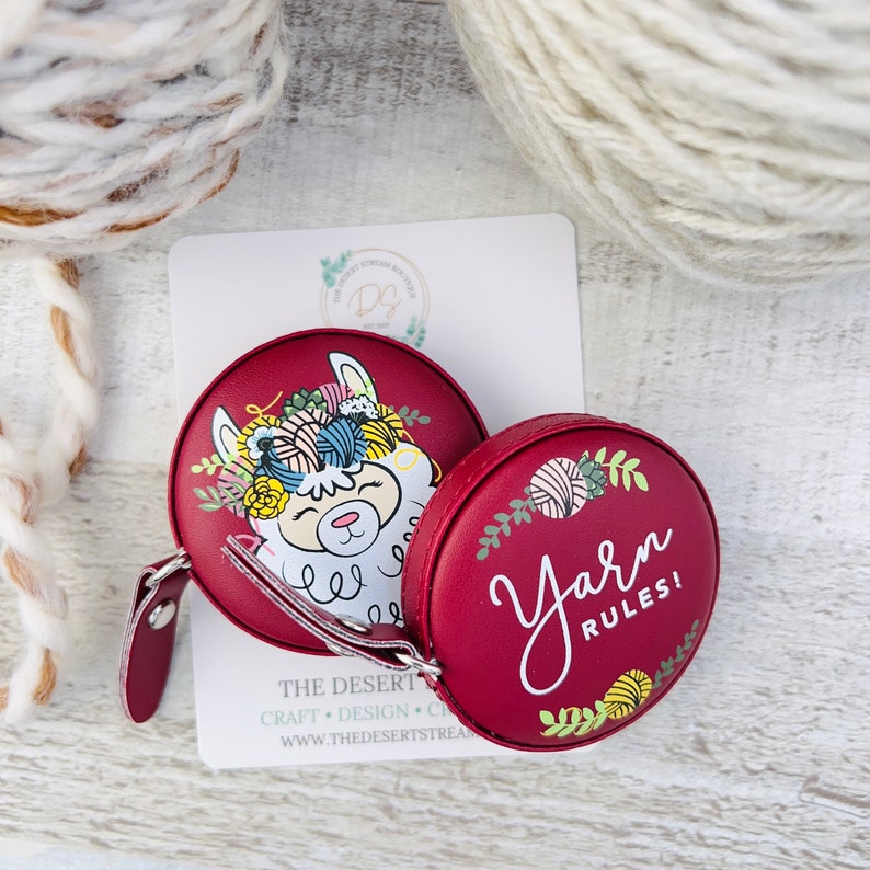 Pu&ograve; includere: Due metri a nastro rotondi bordeaux. Uno presenta un lama con filato e fiori, l'altro dice "Yarn Rules!" in corsivo bianco. Una carta bianca con testo &egrave; dietro i metri a nastro. Matasse di filato sullo sfondo.