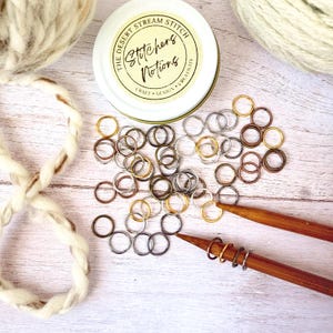 May include: A selection of metal stitch markers in gold, silver, and bronze tones, spread on a light wooden surface. A small tin with the text "The Desert Stream Stitch" and "Stitchers Notions" is visible. Two wooden knitting needles and yarn are also present.