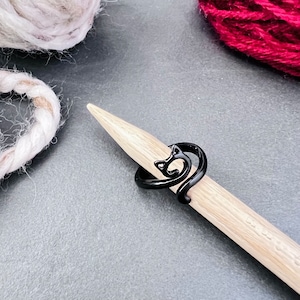 May include: Black cat-shaped ring on a wooden knitting needle. The ring is a simple design with a curled tail and a cat's head.