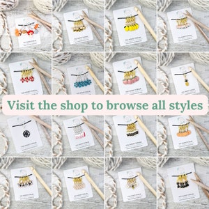 May include: A collection of colorful stitch markers for knitting and crochet, featuring various charms like fruits, animals, and geometric shapes. Each set is displayed on a card with the shop's logo and the text "Visit the shop to browse all styles."