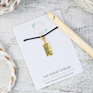 May include: A gold-coloured stitch marker with the words "Find the joy in your journey" engraved on it. The marker is attached to a black cord and is displayed on a white card with the text "The Desert Stream" and website address.