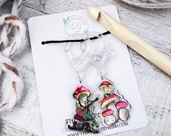 May include: Two clear plastic frog charms with a black cord. The charms feature a frog playing a guitar on a mushroom and a frog sitting on a mushroom. The charms are attached to a white card with the text "The Happy Crochet Stream Boutique" and "www.thehappycrochetstream.com".