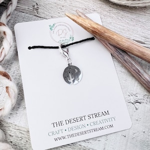 May include: A silver charm with a cactus and moon design on a black cord with a lobster clasp. The charm is on a white card with the text "THE DESERT STREAM" and "CRAFT • DESIGN • CREATIVITY" and "WWW.THEDESERTSTREAM.COM".