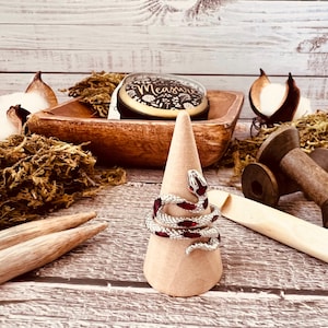 Peut inclure: Une bague en argent en forme de serpent avec des accents en émail rouge sur un présentoir à anneaux conique brun. La bague est entourée d'éléments naturels comme de la mousse, du bois et du coton.