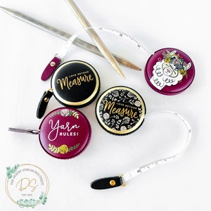 Pu&ograve; includere: Una collezione di metri a nastro rotondi decorativi in nero e bordeaux con dettagli dorati. I nastri presentano frasi come "Love Beyond Measure" e "Yarn Rules!" insieme a disegni floreali e di lama. Sono presenti anche ferri da maglia.