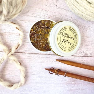 May include: An open white tin of stitch markers with the text "The Desert Stream Stitch, Stitchers Notions, Craft, Design, Creativity". The tin is next to a wooden knitting needle with stitch markers and yarn.