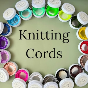 May include: An overhead shot displays multiple small, white tins, each containing colorful knitting cords. The cords are in various shades, including blue, green, yellow, red, and brown. The words "Knitting Cords" are prominently displayed in black text.