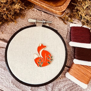 May include: A close-up of a small, round embroidery hoop with a white fabric background. A gold-colored fox pin with green leaves is centered on the fabric. Three spools of embroidery floss are visible in the background.