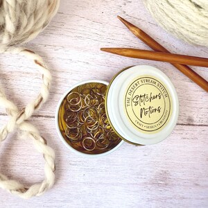 May include: A small, open tin of gold-colored stitch markers next to a partially open tin with the text "Stitchers Notions". Two wooden knitting needles and two balls of yarn are also in the image. The text on the tin reads "The Desert Stream Stitch".