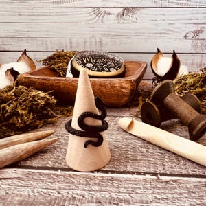 May include: A black snake ring on a cone-shaped ring holder. The ring is made of metal and has a realistic snake design. The ring holder is made of a light brown material and is sitting on a wooden surface. There is a spool of thread, a wooden dowel, and a fabric measuring tape in the background.