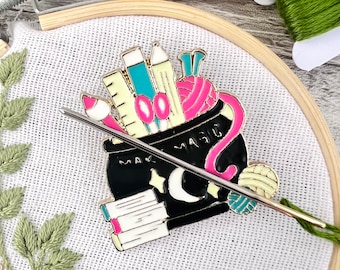 May include: A black cauldron filled with colorful craft supplies, including yarn, pencils, and a ruler. The cauldron has a gold rim and the words "Make Magic" written on the side. A needle threader is placed on top of the cauldron.