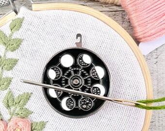 May include: A black and silver moon phase embroidery hoop with a needle threaded through it. The hoop is on a white fabric background with green embroidery.