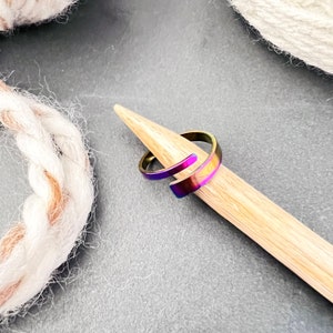 May include: A rainbow iridescent metal ring with a curved design. The ring is resting on a wooden dowel.