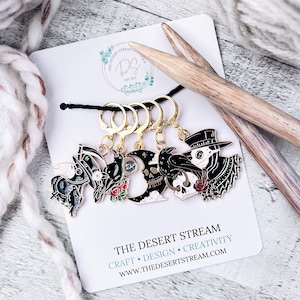 May include: Set of four black and gold metal stitch markers with different spooky Halloween designs. The designs include a skull, a crow, a witch's hat, and a plague doctor. The stitch markers are attached to gold rings. The image also includes a wooden knitting needle and a white yarn ball. The text on the card reads "The Desert Stream Craft Design Creativity www.thedesertstream.com".